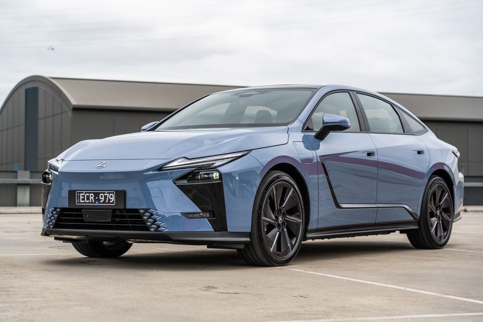 A blue 2027 Lexus ES500e sedan seen from the front-side angle in a car park, with a building in the background
