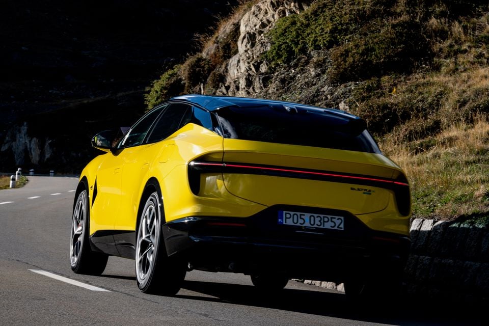 A yellow 2027 Lotus Eletre X SUV with a black roof driving along a mountain road, seen from the rear