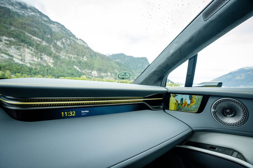 Passenger-side dashboard of the 2027 Lotus Eletre X with a slim digital clock display, yellow trim accents and a door speaker, overlooking mountains