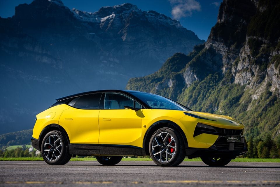 A yellow 2027 Lotus Eletre X electric SUV parked on tarmac in front of towering alpine mountains, seen from the front three-quarter angle