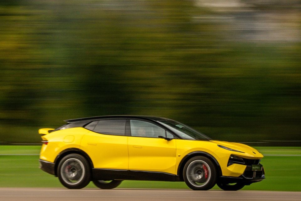 A yellow 2027 Lotus Eletre X electric SUV driving at speed past a blurred green background, seen from the side