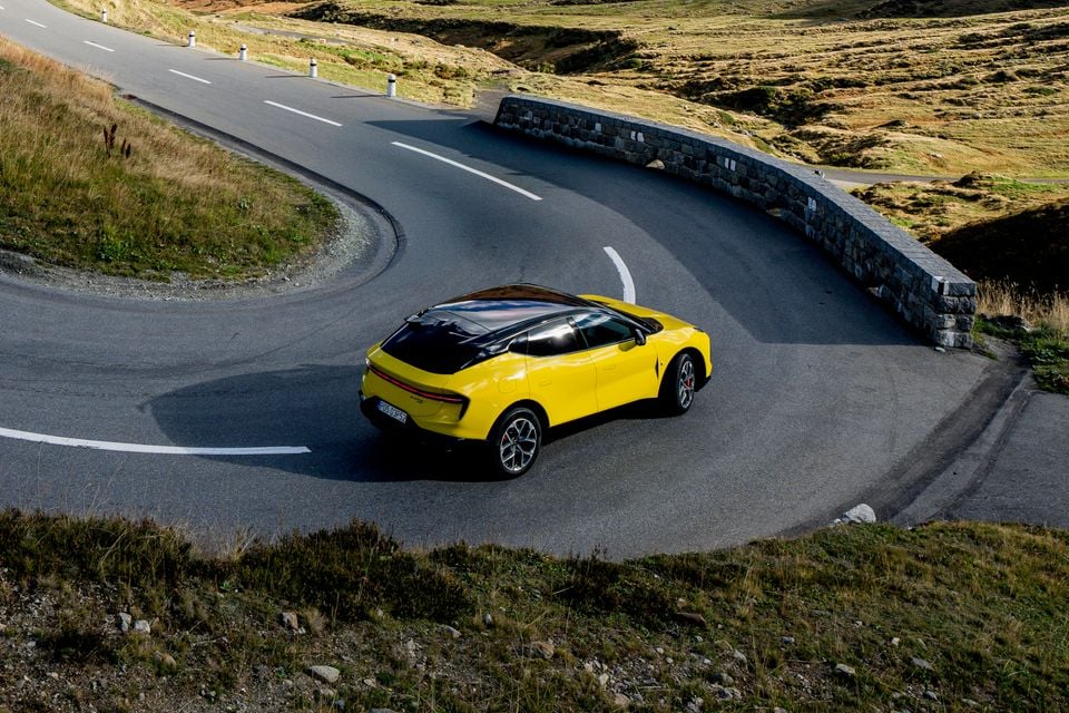 A yellow 2027 Lotus Eletre X SUV seen from the rear, driving around a hairpin bend on a mountain pass beside a stone wall