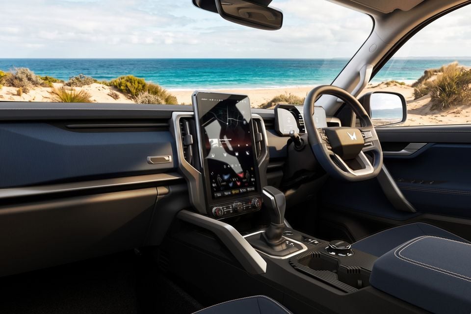 Interior of a Mahindra showing a large portrait-oriented touchscreen infotainment display, steering wheel with Mahindra logo, centre console with gear shifter, and blue upholstery, with a beach visible through the windscreen