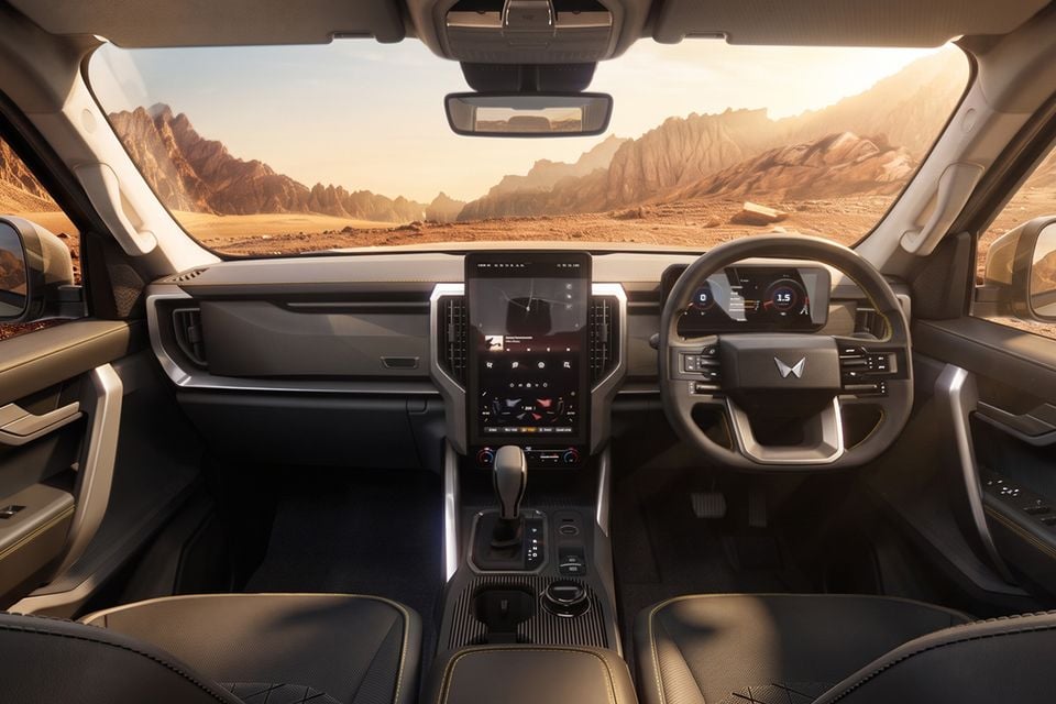Interior of a Mahindra SUV showing the dashboard, large central touchscreen infotainment system, digital instrument cluster, steering wheel, and centre console, with a desert mountain landscape visible through the windscreen.