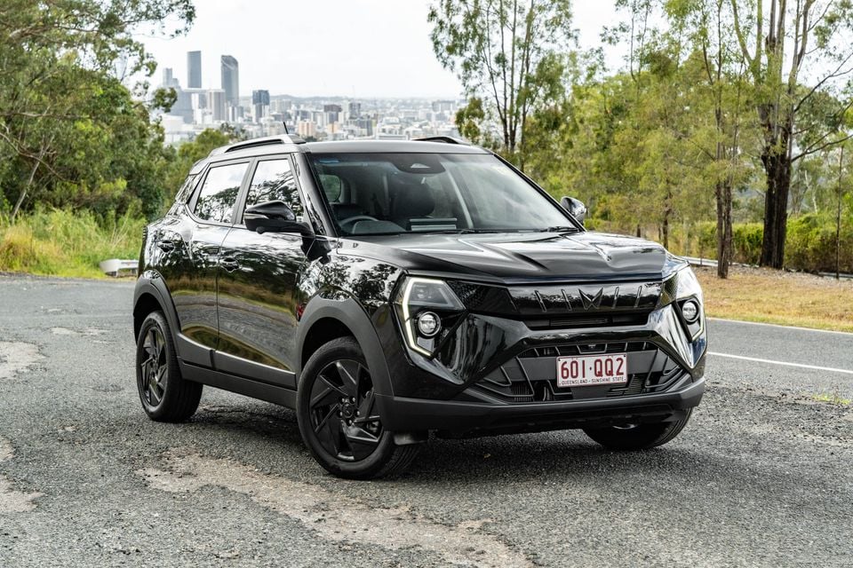 A black Mahindra XUV 3XO Black Edition SUV seen from the front-side angle, parked on a road overlooking Brisbane's city skyline with trees in the background.