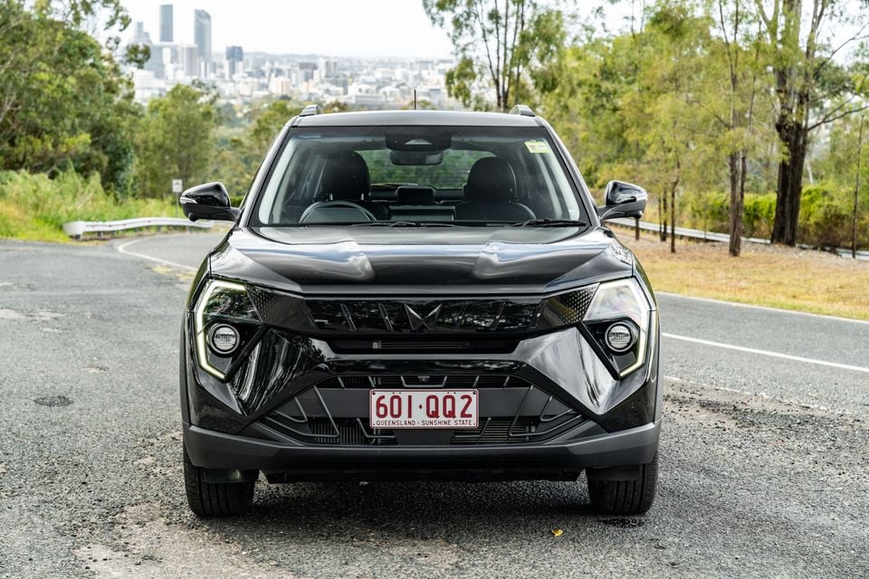 A black Mahindra XUV 3XO SUV seen from the front, parked on a road with Brisbane's skyline visible in the background.