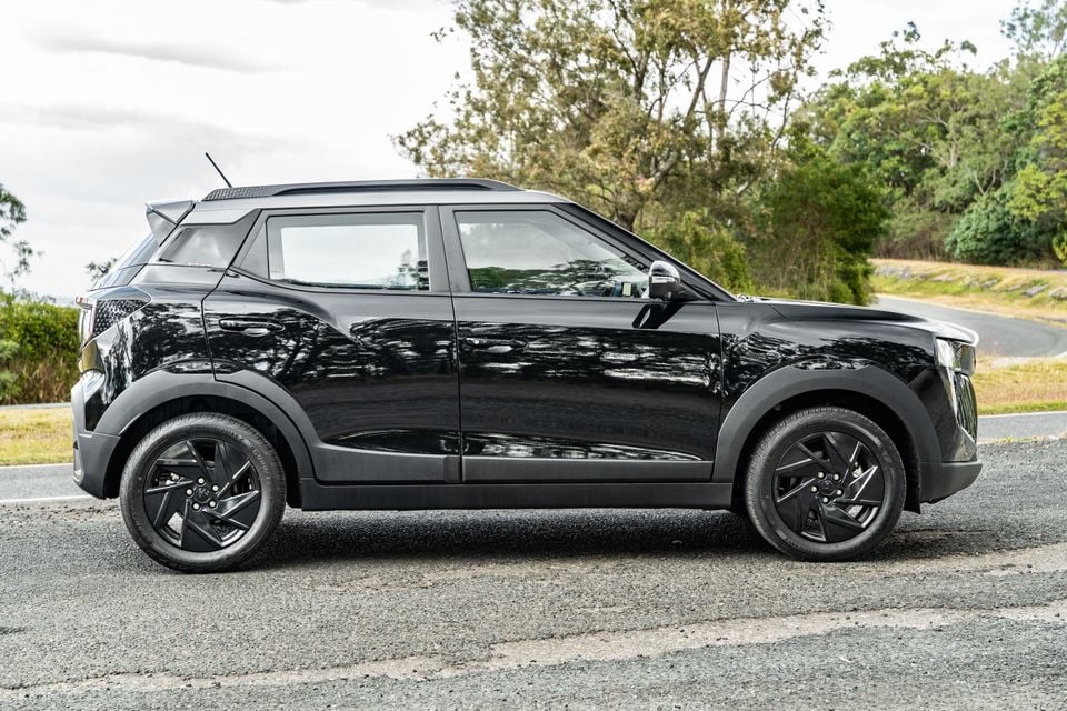 A black Mahindra XUV 3XO small SUV seen from the side, parked on a road with trees in the background.