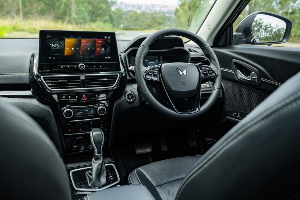 Interior of a Mahindra XUV 3XO showing the dashboard, touchscreen infotainment system, steering wheel with Mahindra logo, dual-zone climate controls, and manual gear shifter from the driver's side