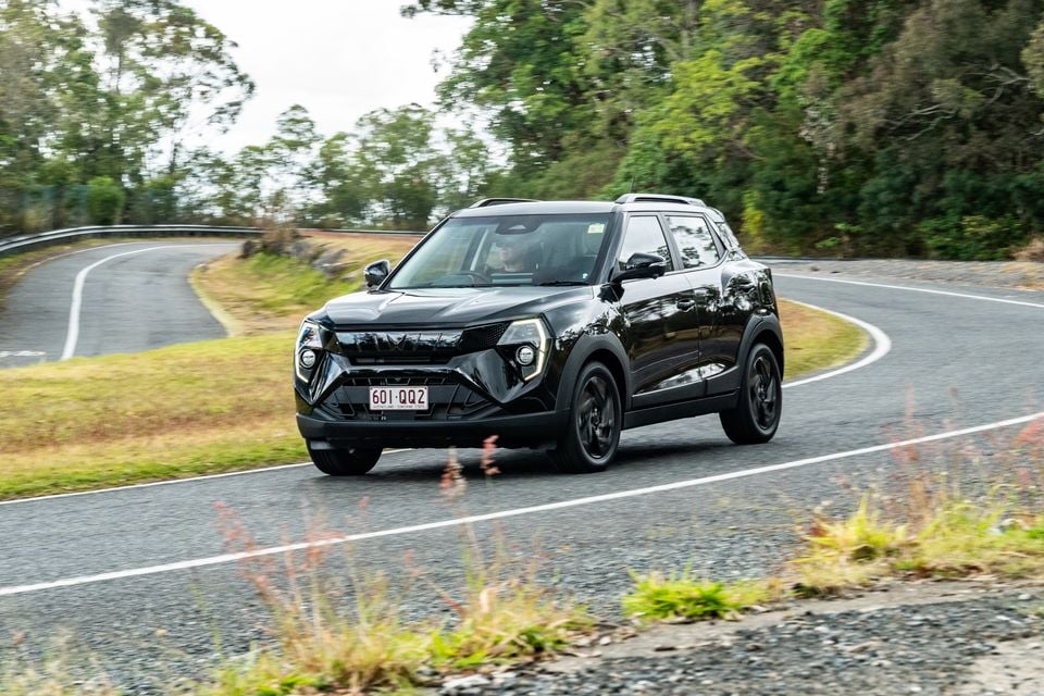 A black Mahindra XUV 3XO SUV driving along a winding road surrounded by trees, seen from the front-side