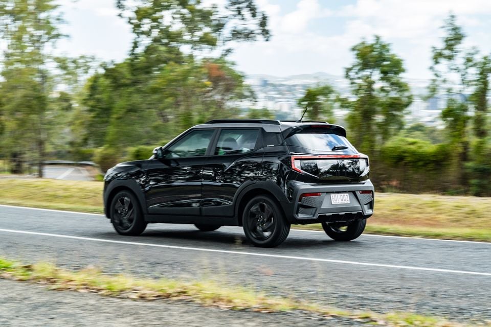 A black Mahindra XUV 3XO Black Edition small SUV driving on a road, seen from the rear three-quarter angle, with trees and hills in the background.
