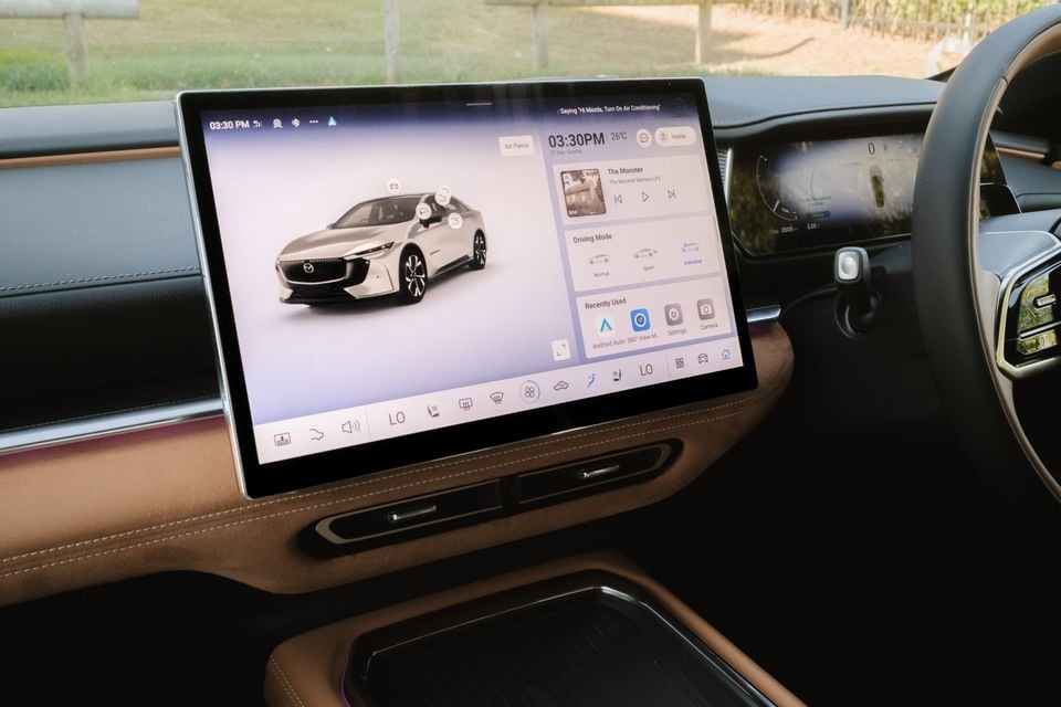 The large infotainment touchscreen in a Mazda 6e Atenza shows a vehicle status display with driving mode and media controls, seen from the driver's seat.