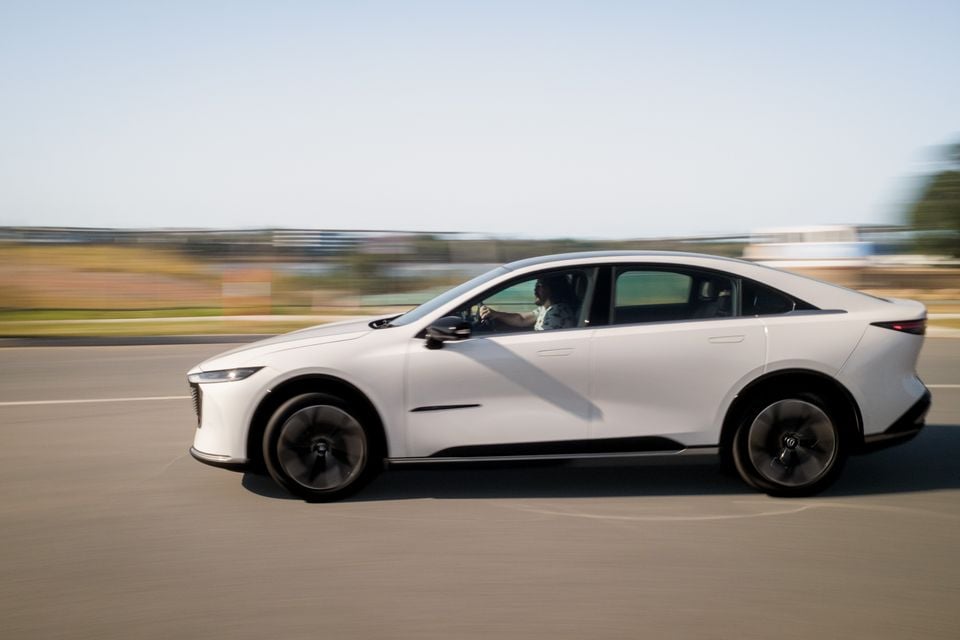 A white 2027 Mazda 6e Atenza sedan photographed from the side while driving on a road