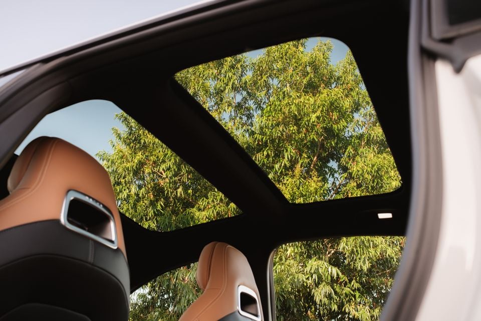 A large panoramic sunroof lets in a view of tree branches above the tan leather front seats of a 2027 Mazda 6e Atenza.