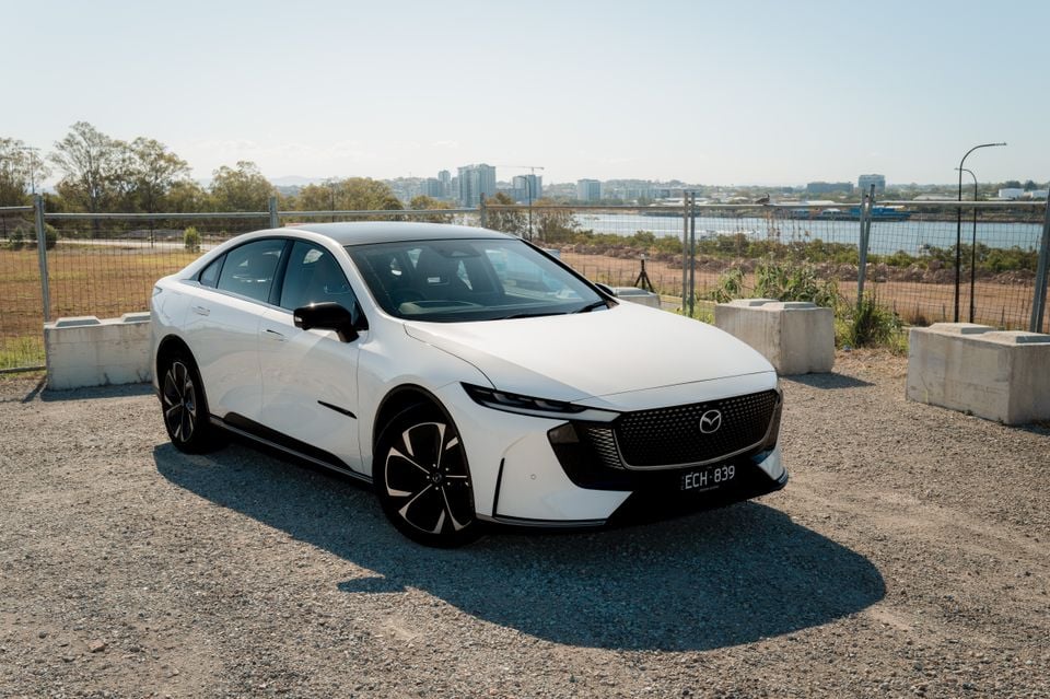 A white 2027 Mazda 6e Atenza sedan parked on gravel with a city skyline and harbour in the background.