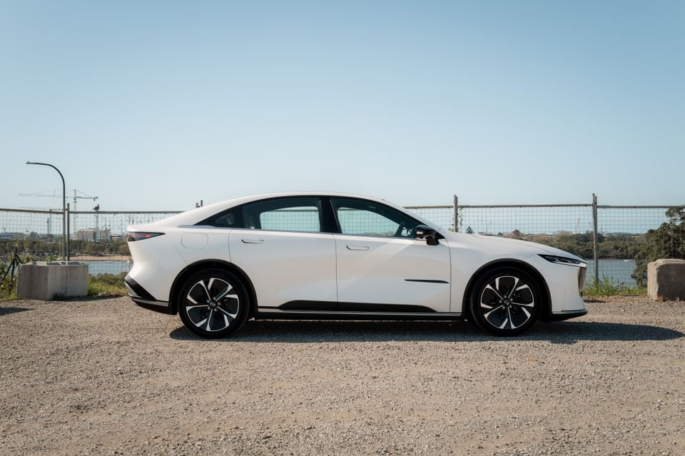 A white 2027 Mazda 6e Atenza sedan parked on gravel, seen from the side, with a river and city skyline in the background.