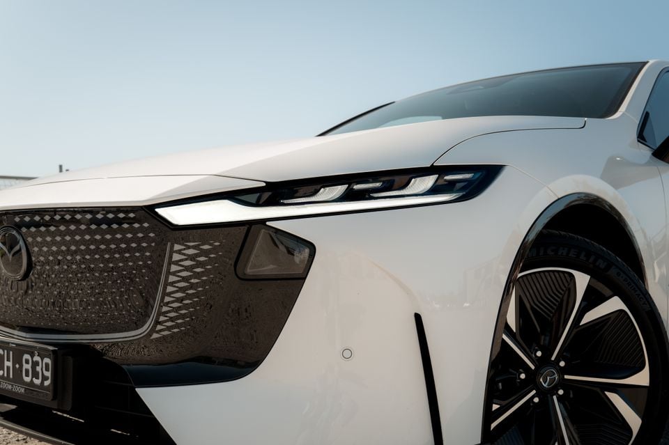 Close-up of the front headlight, grille and wheel of a white 2027 Mazda 6e Atenza sedan.