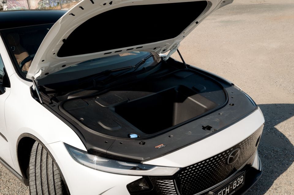 The white 2027 Mazda 6e Atenza is shown with its front bonnet open, revealing an empty front storage compartment (frunk) instead of an engine.