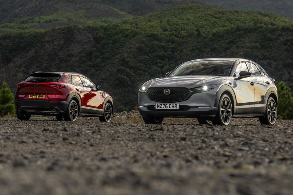 A grey 2027 Mazda CX-30 seen from the front beside a red CX-30 seen from the rear, parked on gravel with forested hills behind