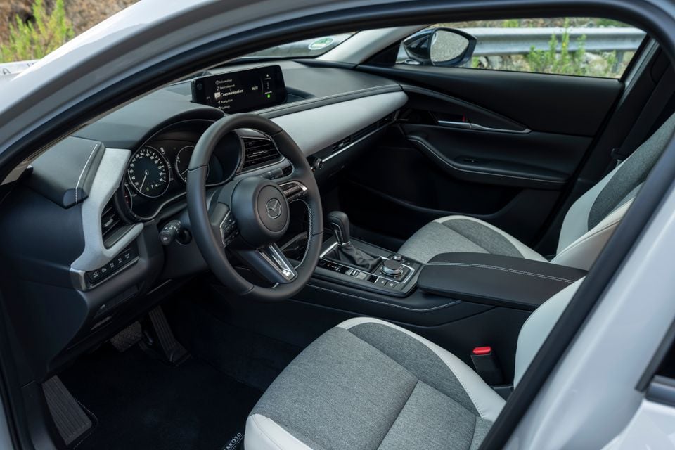 Interior of a 2027 Mazda CX-30 seen through the open driver's door, showing the steering wheel, digital instruments, infotainment screen and grey cloth front seats