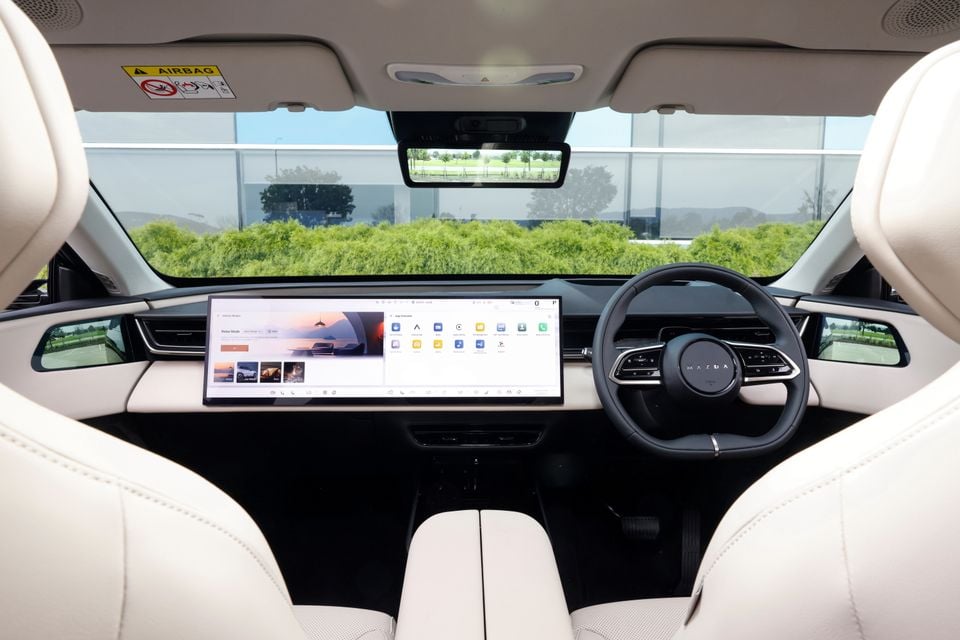 The interior of a 2027 Mazda CX-6e shows a wide dashboard touchscreen, steering wheel and cream leather seats viewed from the back seat.