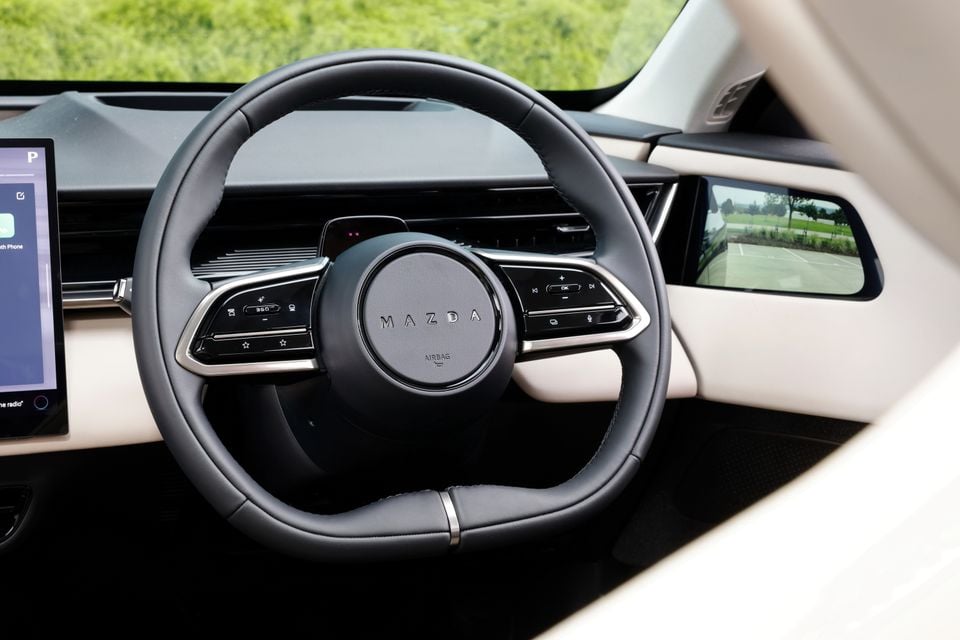 Close-up view of the steering wheel and touchscreen in a 2027 Mazda CX-6e, showing the digital instrument display and side mirror camera monitor