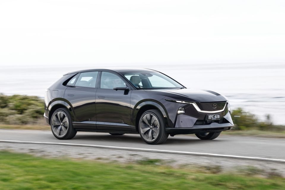A dark 2027 Mazda CX-6e electric SUV driving along a coastal road, seen from the front quarter.
