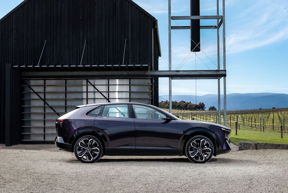 A dark purple 2027 Mazda CX-6e SUV parked in profile outside a black barn, with vineyards visible in the background.