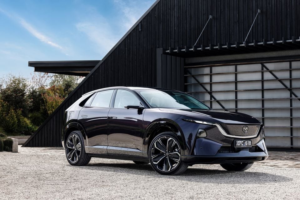 A dark purple 2027 Mazda CX-6e electric SUV parked outside a modern black timber building, seen from the front quarter view.