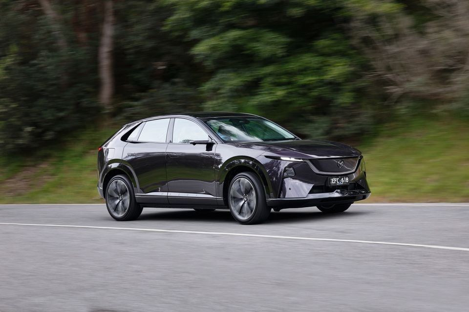 A dark purple 2027 Mazda CX-6e SUV driving along a road, seen from the front quarter view