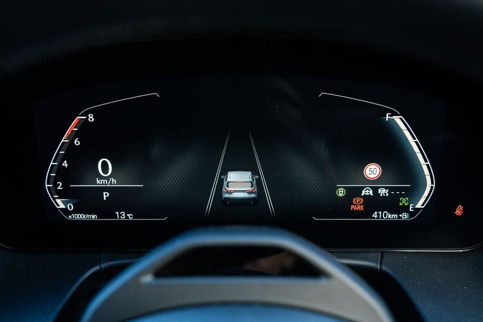 Digital instrument cluster in a Mazda CX-5 showing speed, RPM, temperature, fuel level, and driver-assist icons viewed from behind the steering wheel
