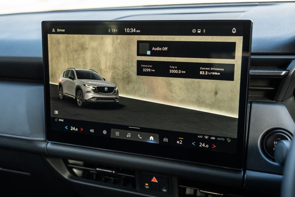 The infotainment touchscreen of a Mazda CX-5 showing the home screen with odometer, trip and efficiency data, climate controls, and an image of the vehicle