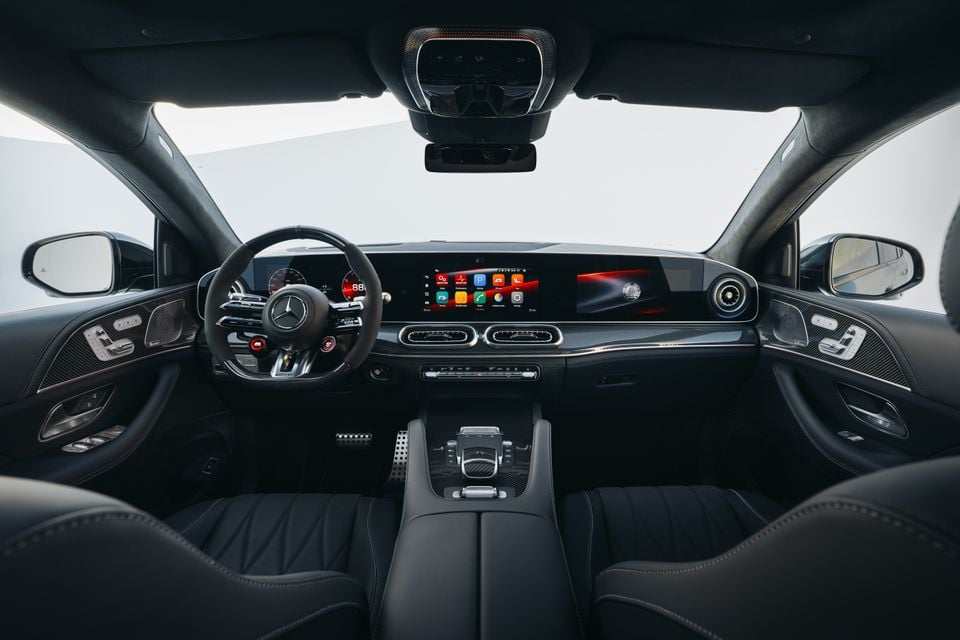 Interior of a Mercedes-AMG GLC 63 S Coupe showing the dashboard, AMG flat-bottom steering wheel, wide infotainment display with touchscreen, carbon fibre trim, and black leather seats
