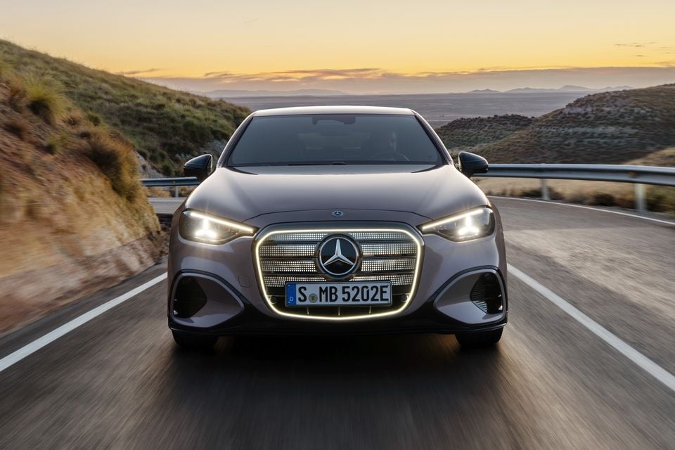 Um sedã Mercedes-Benz Classe C elétrico 2027 com grade iluminada dirigindo em uma estrada sinuosa na encosta ao pôr do sol, visto de frente
