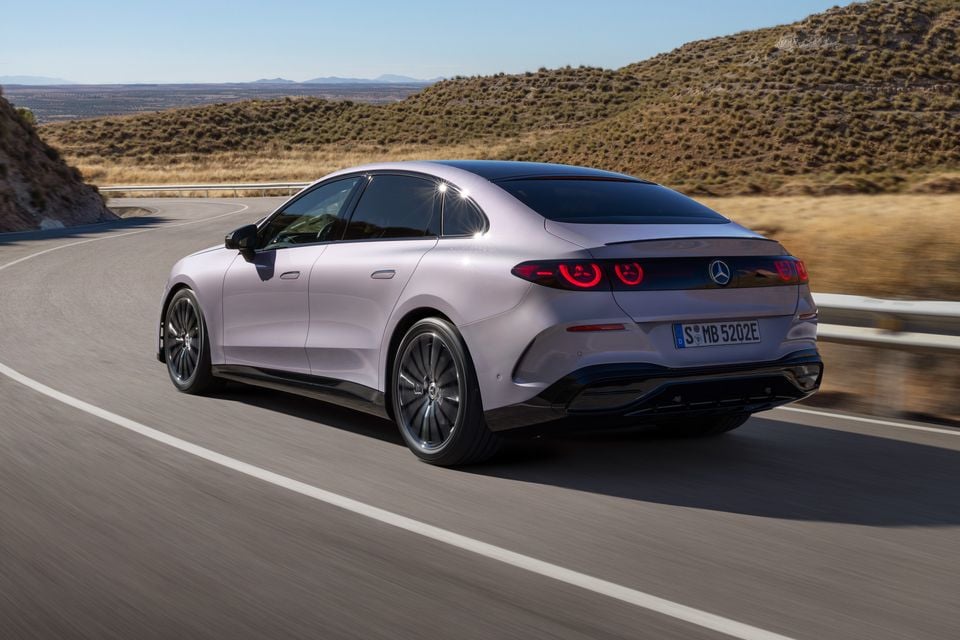 A silver-lilac 2027 Mercedes-Benz C-Class Electric sedan driving on a winding hillside road, seen from the rear