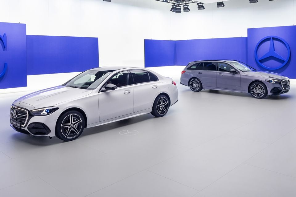 A white Mercedes-Benz C-Class sedan and a grey C-Class wagon displayed together in a studio, with the Mercedes-Benz logo visible on the wall behind them.