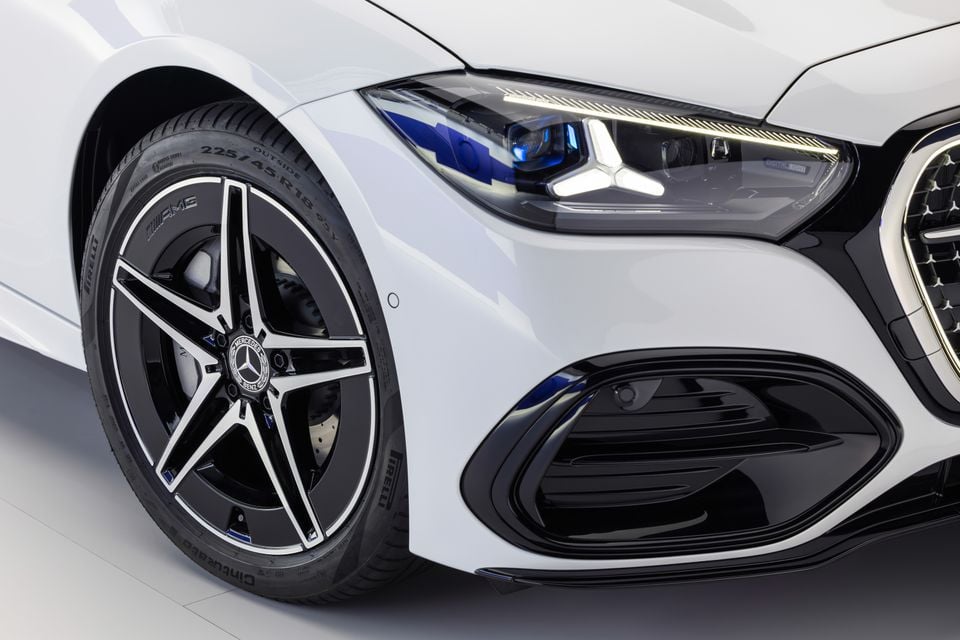 Close-up of the front corner of a white Mercedes-Benz C-Class facelift, showing the AMG-style 18-inch alloy wheel with Pirelli tyre and illuminated multibeam headlight