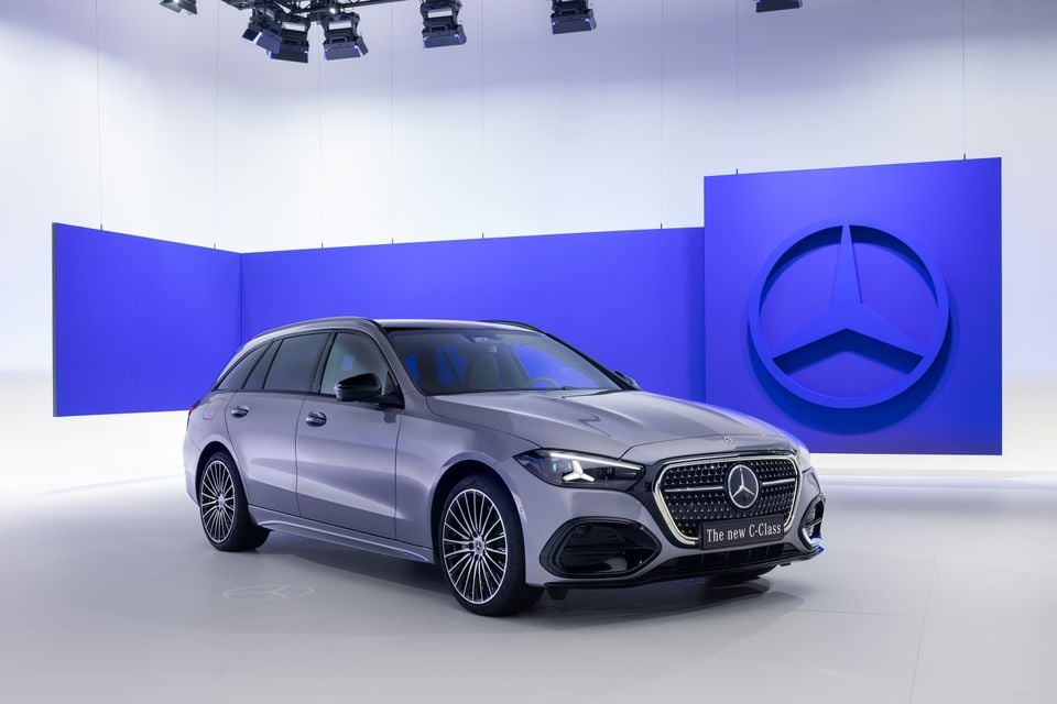 A grey Mercedes-Benz C-Class wagon seen from the front-left in a studio setting, with a large Mercedes-Benz star logo on a blue backdrop behind it.