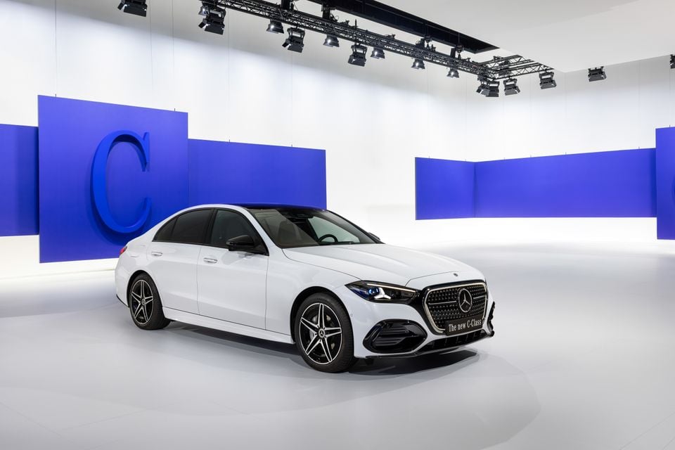 A white 2027 Mercedes-Benz C-Class sedan seen from the front-side angle at a studio reveal event, with blue branded backdrop panels behind it.