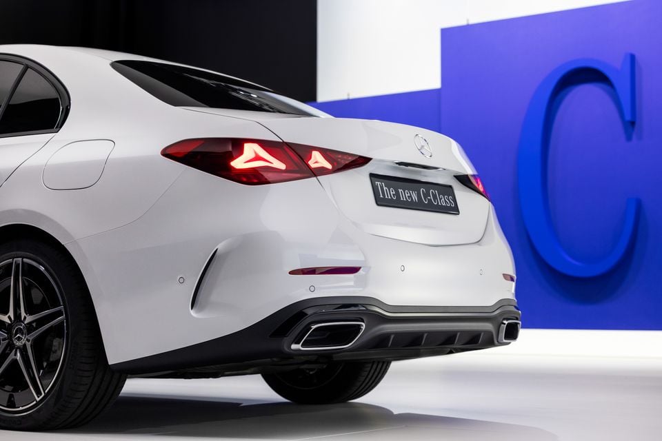Rear view of a white Mercedes-Benz C-Class sedan at a reveal event, showing tail lights, dual exhausts, and a badge reading 'The new C-Class'