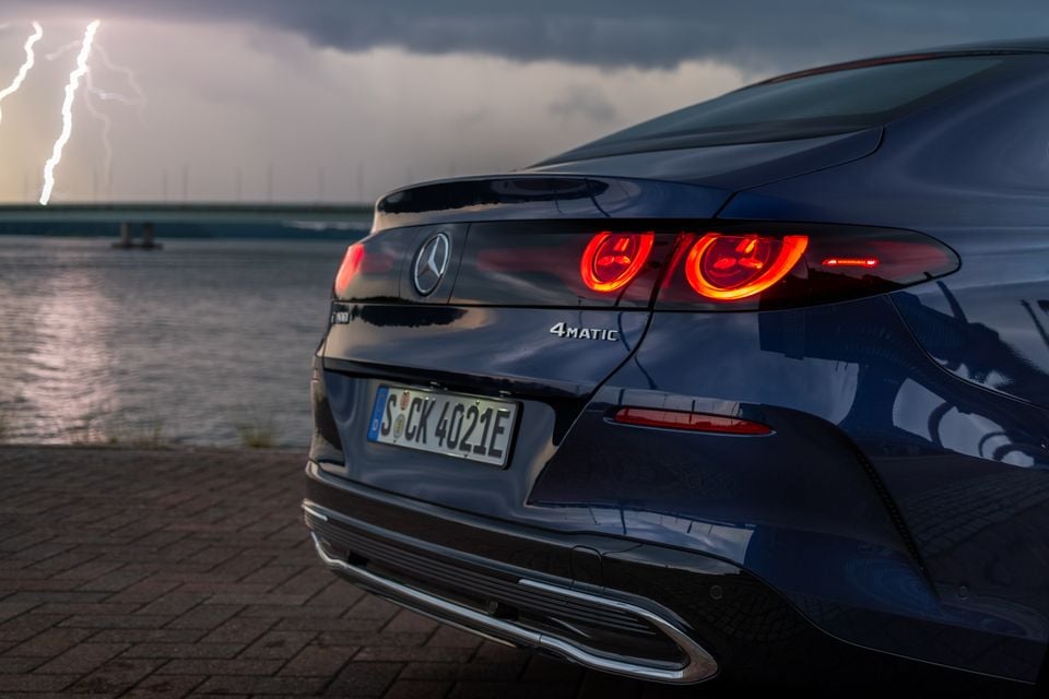Rear close-up of a dark blue 2027 Mercedes-Benz C-Class Electric 4MATIC sedan, showing distinctive circular tail lights illuminated against a stormy sky with lightning over water