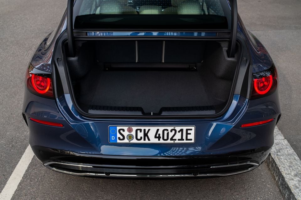 Open boot of a dark blue 2027 Mercedes-Benz C-Class electric sedan, seen from above the rear, showing a spacious empty cargo area with carpeted floor and round red tail lights either side