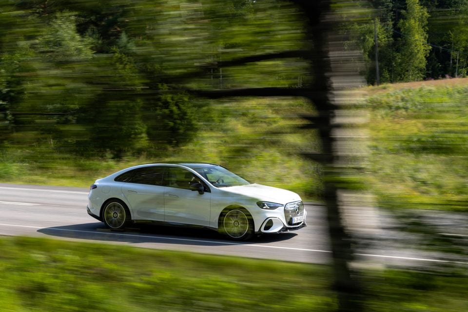 A white Mercedes-Benz C-Class electric sedan driving along a tree-lined road, seen from the side with motion blur conveying speed.