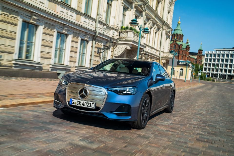 A blue Mercedes-Benz C-Class electric sedan driving along a cobblestone street past grand European architecture