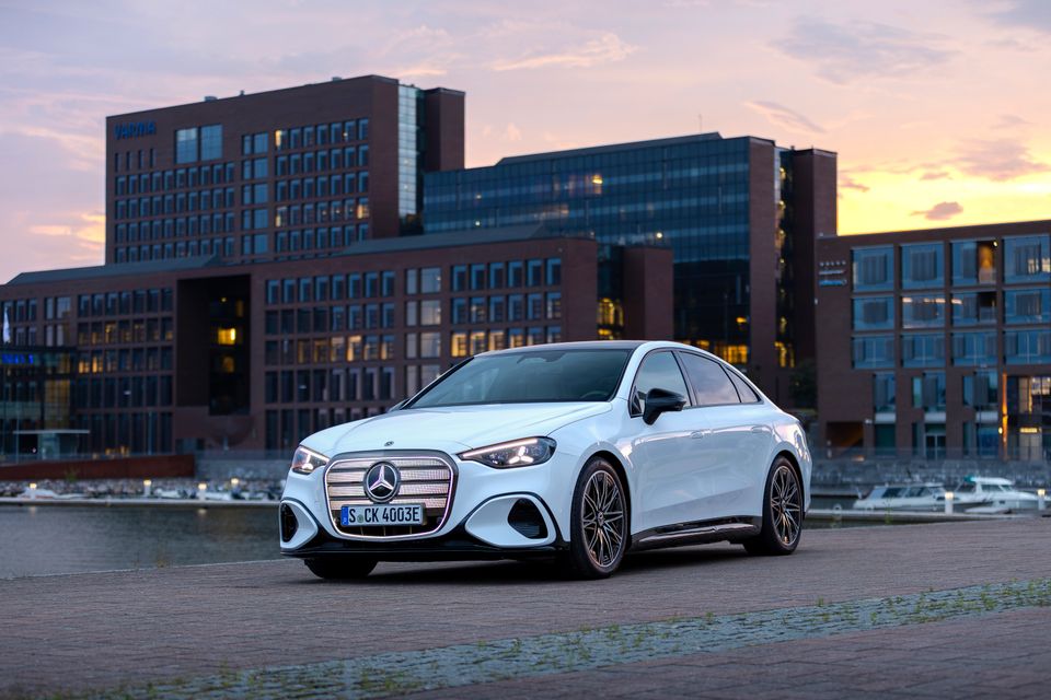 A white 2027 Mercedes-Benz C-Class electric sedan seen from the front-side, parked on cobblestones near a waterfront at sunset with modern buildings in the background.