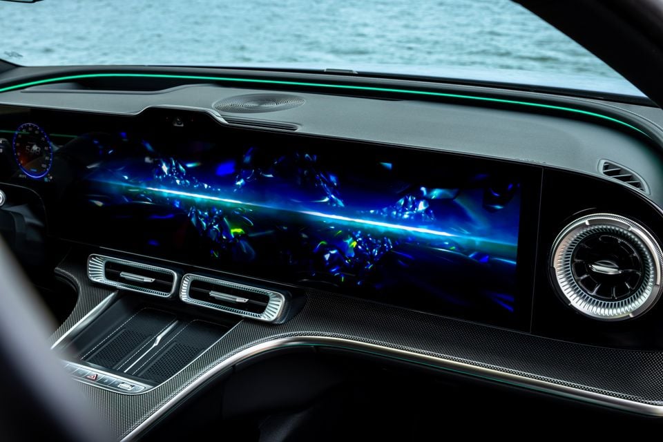 Interior dashboard of a 2027 Mercedes-Benz C-Class Electric showing a large widescreen infotainment display with a vibrant blue animated wallpaper, ambient lighting, and carbon fibre trim