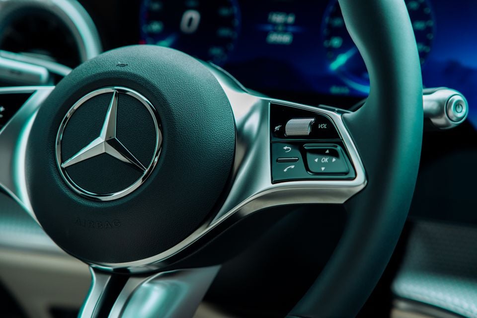 Close-up of a Mercedes-Benz steering wheel showing the three-pointed star logo, multifunction controls, and digital instrument cluster in the background