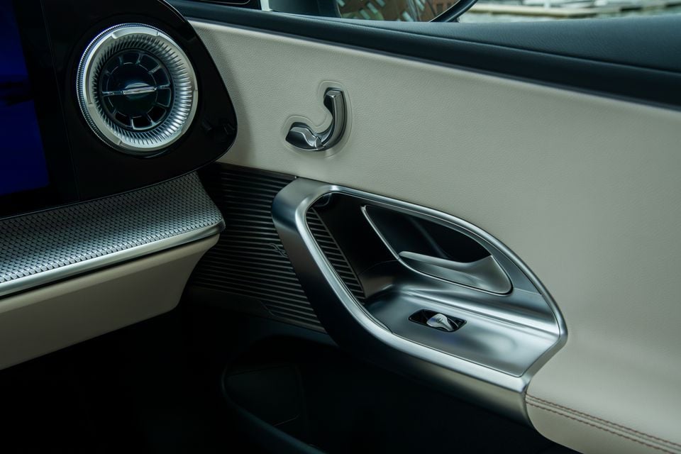 Close-up of the passenger door panel interior of a 2027 Mercedes-Benz C-Class electric, showing a circular air vent, a coat hook, door handle recess, and premium cream leather trim with contrast stitching.
