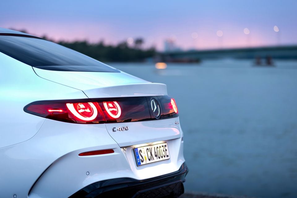 Close-up of the rear of a white Mercedes-Benz C400 electric sedan, showing illuminated tail lights and badge at dusk near a waterway