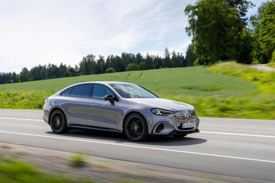 A grey Mercedes-Benz C-Class electric sedan driving along a country road, seen from the front-side angle with green fields in the background.