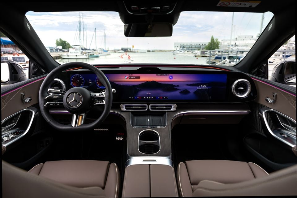 Interior of a 2027 Mercedes-Benz C-Class Electric showing the wide curved touchscreen display, steering wheel, and tan leather seats, parked near a marina